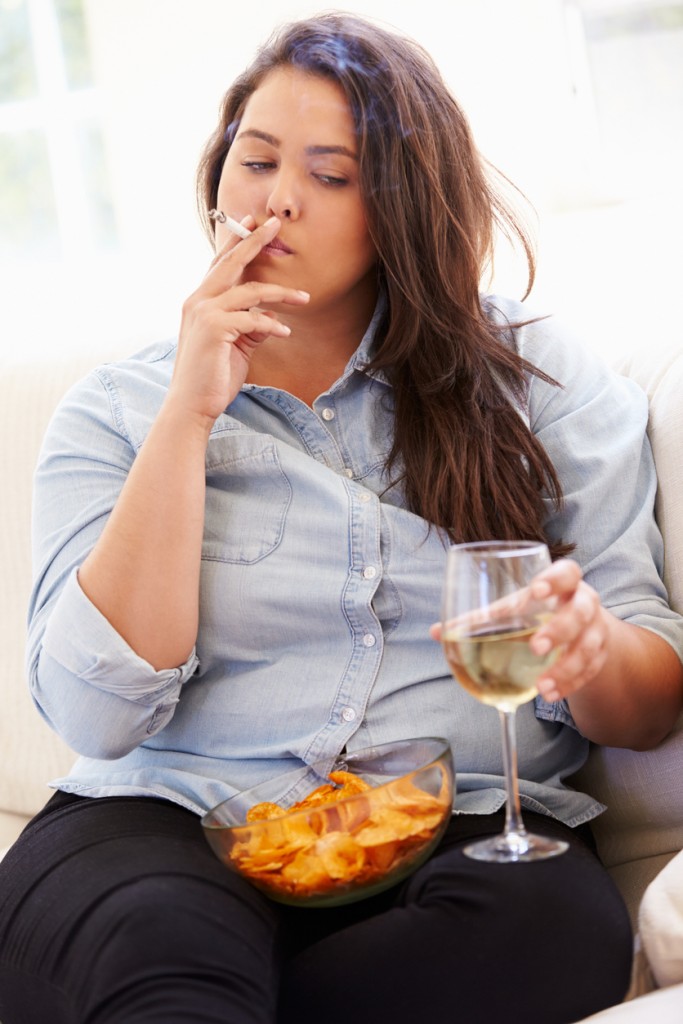 Overweight Woman Eating Chips, Drinking Wine And Smoking Whilst At Home Sitting On Sofa