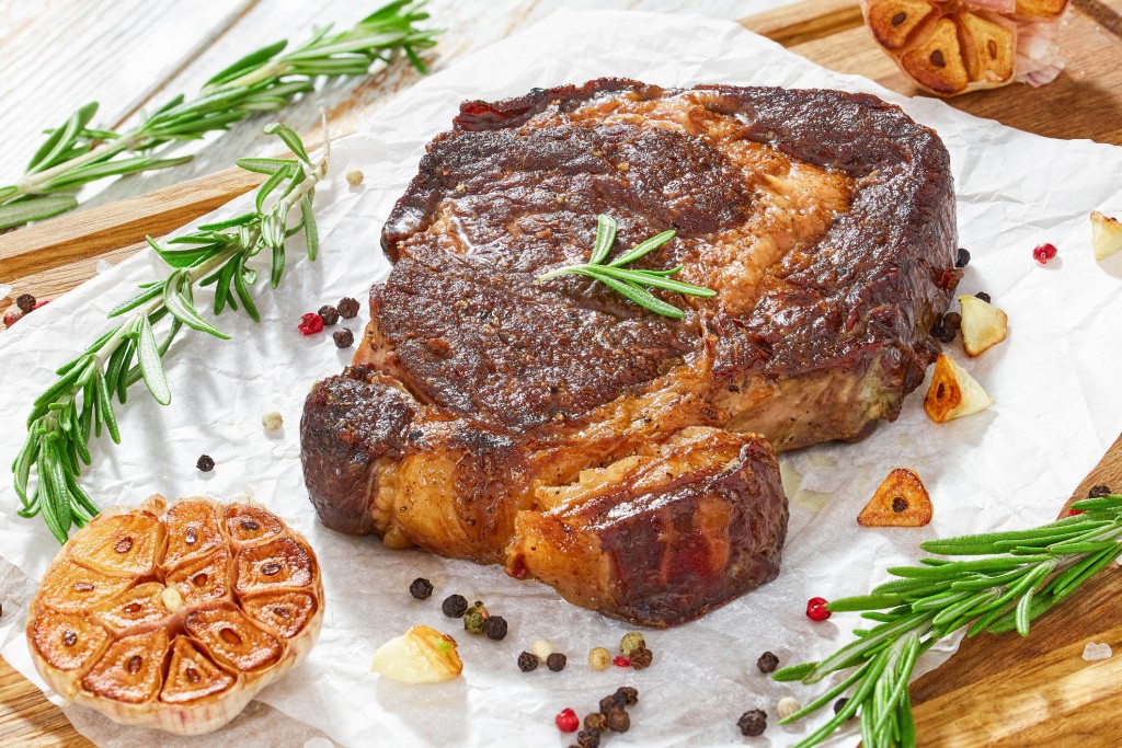 medium-rare grilled rib-eye steak with garlic and rosemary on paper on wooden background, close-up