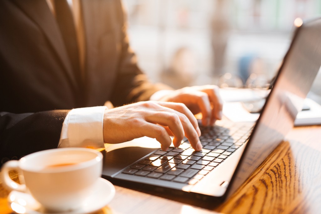 Successful businessman wearing black formal suit working by window in rays of golden sunset light: closeup shot of male hands typing at laptop against background of coffee cup and business tools