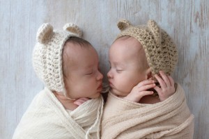Profile headshot of two, fraternal, twin, baby girls sleeping. They are wearing crocheted bear hats and are swaddled in cream and tan wraps.