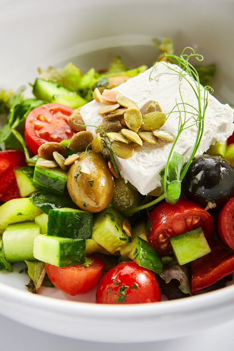Greek salad in white bowl. Sliced fresh vegetables and feta cheese. Served traditional Greece cuisine. Cut raw tomatoes, cucumbers with olive oil in plate. Restaurant food portion close up