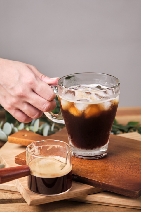 woman used hand take cold iced black brew coffee in a glass with pieces of ice on a wooden table, natural light, fresh summer drink in the morning