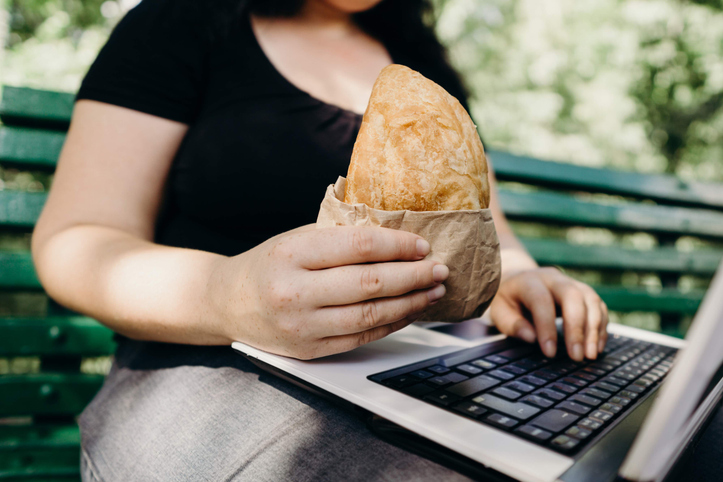 Unhealthy fast food,high-calorie snack, sedentary working, eating on the go. Close up of overweight woman with fattening take-out pastry typing on laptop in the street
