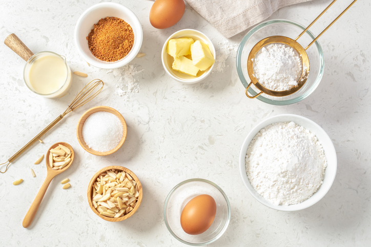 Baking and cake ingredients in bowls, butter, milk, nuts and eggs with gold flour sieve and whisk on marble background, flatlay