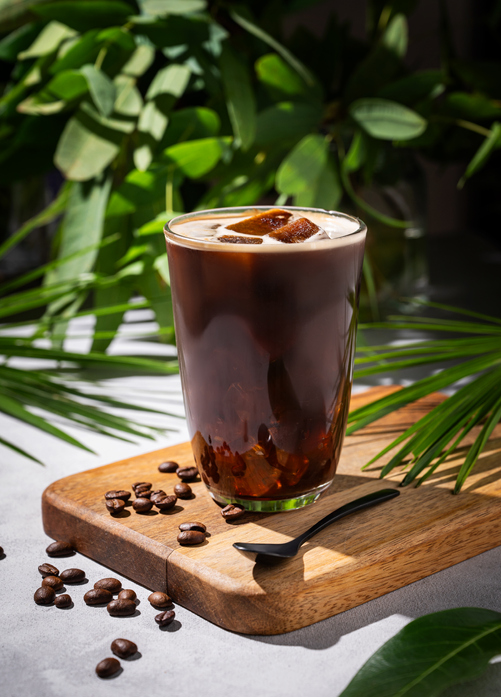 Iced coffee brew in a glass on a wooden board on a light background with coffee beans and morning shadows in tropical branches. Summer refreshment concept.